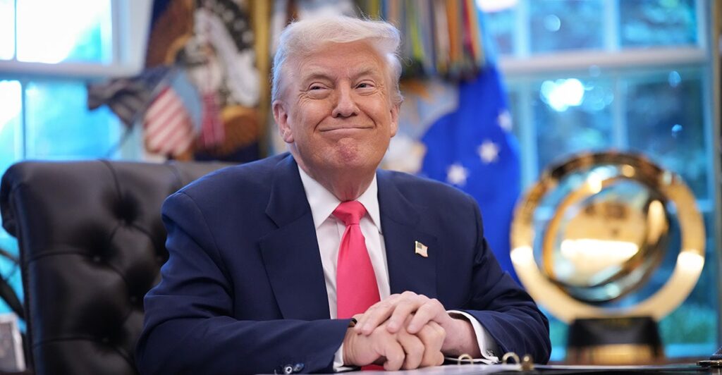 Donald Trump sitting at his oval office desk with hands folded and smiling