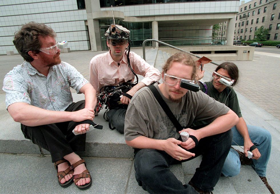 Rehmi Post, Thad Starner, Steve Mann and Professor Alex Pentland donning wearable computers that resemble electronic eyeglasses in the 1990s. They are sitting on steps outside of a building at MIT.