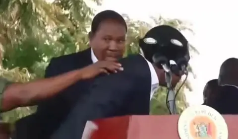 X President Hakainde Hichilema crouches behind a microphone. A hand can be seen reaching out from the left hand side of the frame touching the president on the back. A man behind the president is looking nervous.