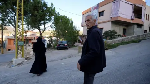 Neha Sharma/BBC An older man is seen on a road, with a woman nearby, in front of a building which has a large image on the side.