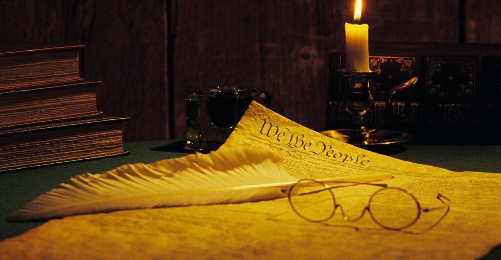 Spectacles and quill pen lay atop a copy of the U.S. Constitution next to a lit candle.