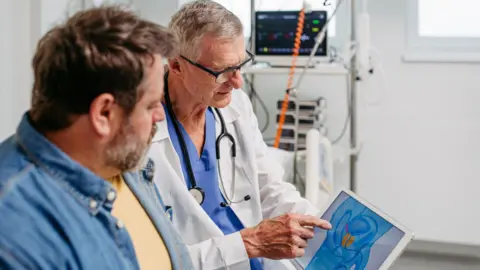 Getty Images A doctor wearing a white coat and a stethoscope around his neck looks at a tablet showing the location of the prostate, with a male patient next to him. The  patient is wearing a denim shirt and has short, brown hair and a greying beard.