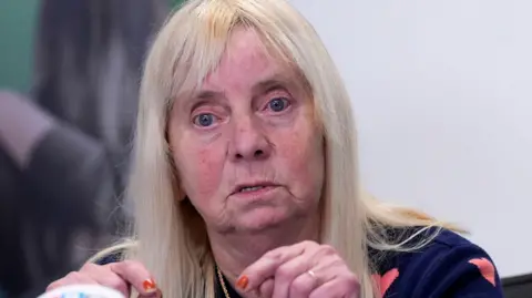 PA Media Margaret Aspinall with long blond hair wearing a navy jumper with peach hearts on it speaking at a press conference.