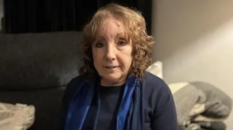 An older woman with pale curly hair sits on a grey sofa smiling. 