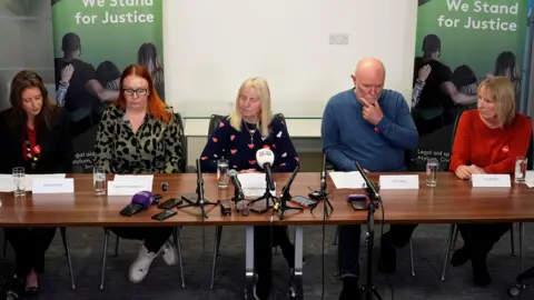 Left to right: Nicola Brook, Charlotte Hennessy, Margaret Aspinall, Steve Kelly and Sue Roberts at the press conference. There are sitting behind a table with posters behind them saying 'We Stand for Justice'.