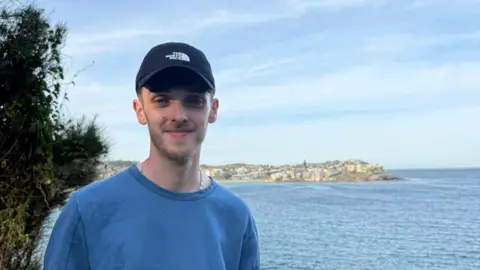 Kian Bentley Kian Bentley is wearing a black cap and a blue T-shirt and is looking straight at the camera. The blue sea is behind him along with a headland