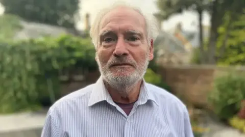 Sammy Stein looking straight at the camera. He has white hair and a grey and white beard. He is wearing a white shirt with blue pinstripes.