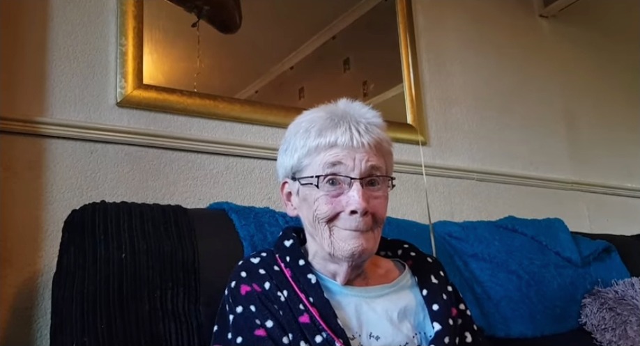 An elderly woman with short white hair and glasses smiles while wearing a dark robe with white and pink hearts.