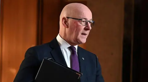 Getty Images John Swinney looking slightly off camera. He is wearing a dark suit over a white shirt with a dark tie