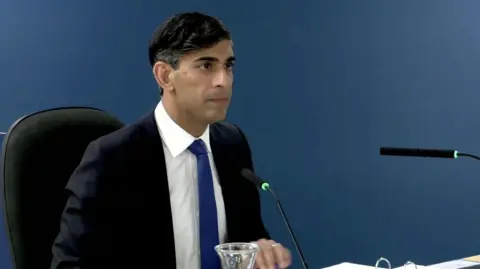 PA Media Rishi Sunak sitting down at the Covid inquiry giving evidence. He is wearing a dark-blue suit with a white shirt and royal blue tie. 