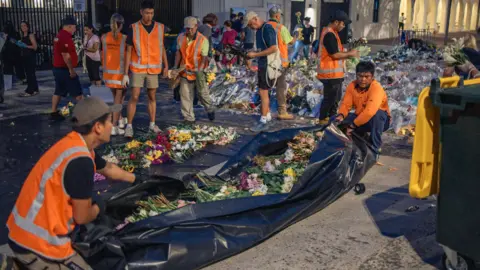 Instagram/@picciesforpotato Workers in high-vis clothing wrap up floral tributes in massive black sheets of plastic