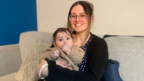 Chloe with her brown hair tied back, looks into the camera smiling. She is wearing a black cardigan over a black and white patterned dress. She is cuddling her six-month-old baby who has her fist in her month and is wearing a knitted, beige cardigan. They are sat on a grey couch with a navy cushion behind her.