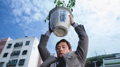 CJ Entertainment Lee Byung-hun in No Other Choice, holding a plant pot above his head