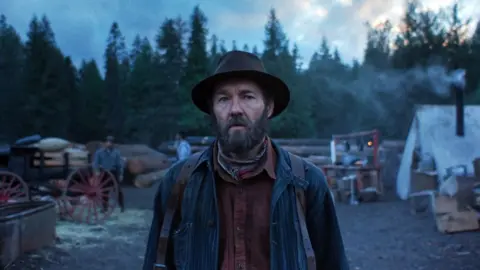 Netflix Joel Edgertonin Train Dreams, wearing a hat and looking ahead, with trees in the background