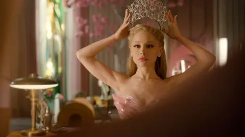 Universal Ariana Grande in Wicked: For Good, as Glinda the good witch, wearing a pink dress and placing a tiara on her head