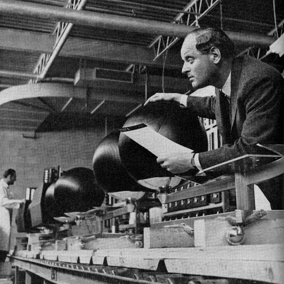 Black and white photo of a line of stereo components under assembly, with a man in a lab coat at one end and a man in a suit at the other.  