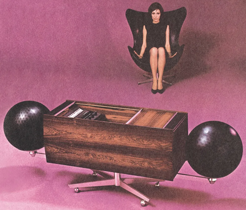 Color photo of a modern stereo system in the foreground and a woman sitting in a modern arm chair in the back. 