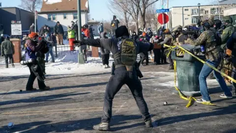 EPA A hooded federal officer with their back to the shot with their arms outstretched about to throw what appears to be a tear gas canister as protesters look on from snowy porches and other federal agents are behind the officer.