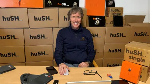 Hush Paul Nugent sitting at a wooden desk with piles of cardboard boxes printed with Hush on them behind him.