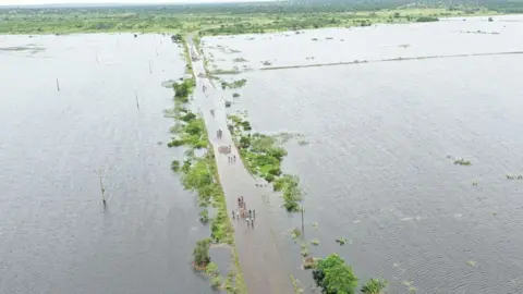 AFP/Getty Images Aerial view showing people residents wading through floodwater to cross a road near Maputo on January 20, 2026.