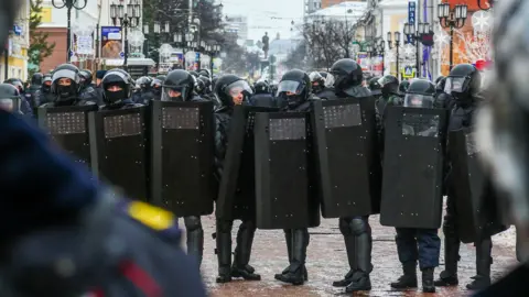 Aleksey Fokin/SOPA Images/LightRocket via Getty Images A row of police officers, wearing black uniforms with riot shields, helmets and protective leg gear, stretches across a street in a town centre. 