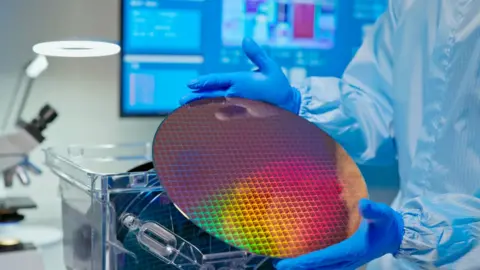 Getty Images A technician holds up a silicon wafer - a round flat disc reflecting pink, red, yellow and green colours. 