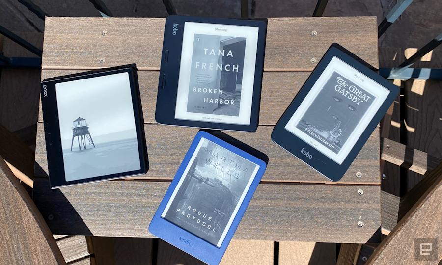 Four ereaders are arranged on a dark brown wooden table outside. There are Boox, Kindle and Kobo devices showing the covers of different novels from Tana French, F. Scott Fitzgerald and Martha Wells.  
