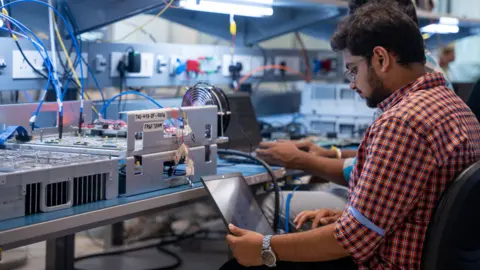 Tejas Networks A man in a checked shirt looks at his laptop, while sitting at a workbench where electrical equipment is being worked on.