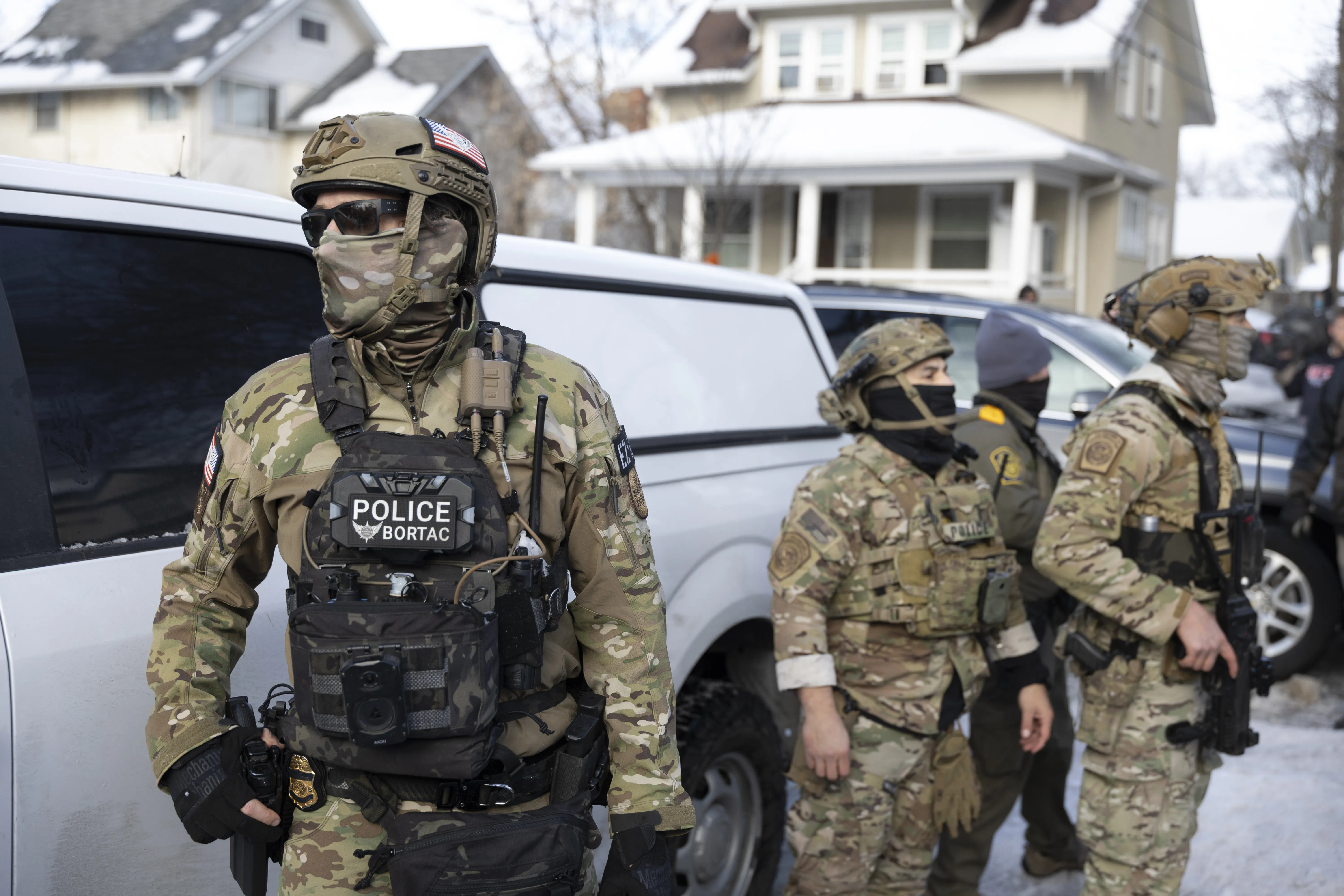 ICE agents in paramilitary-style gear in front of vehicles.