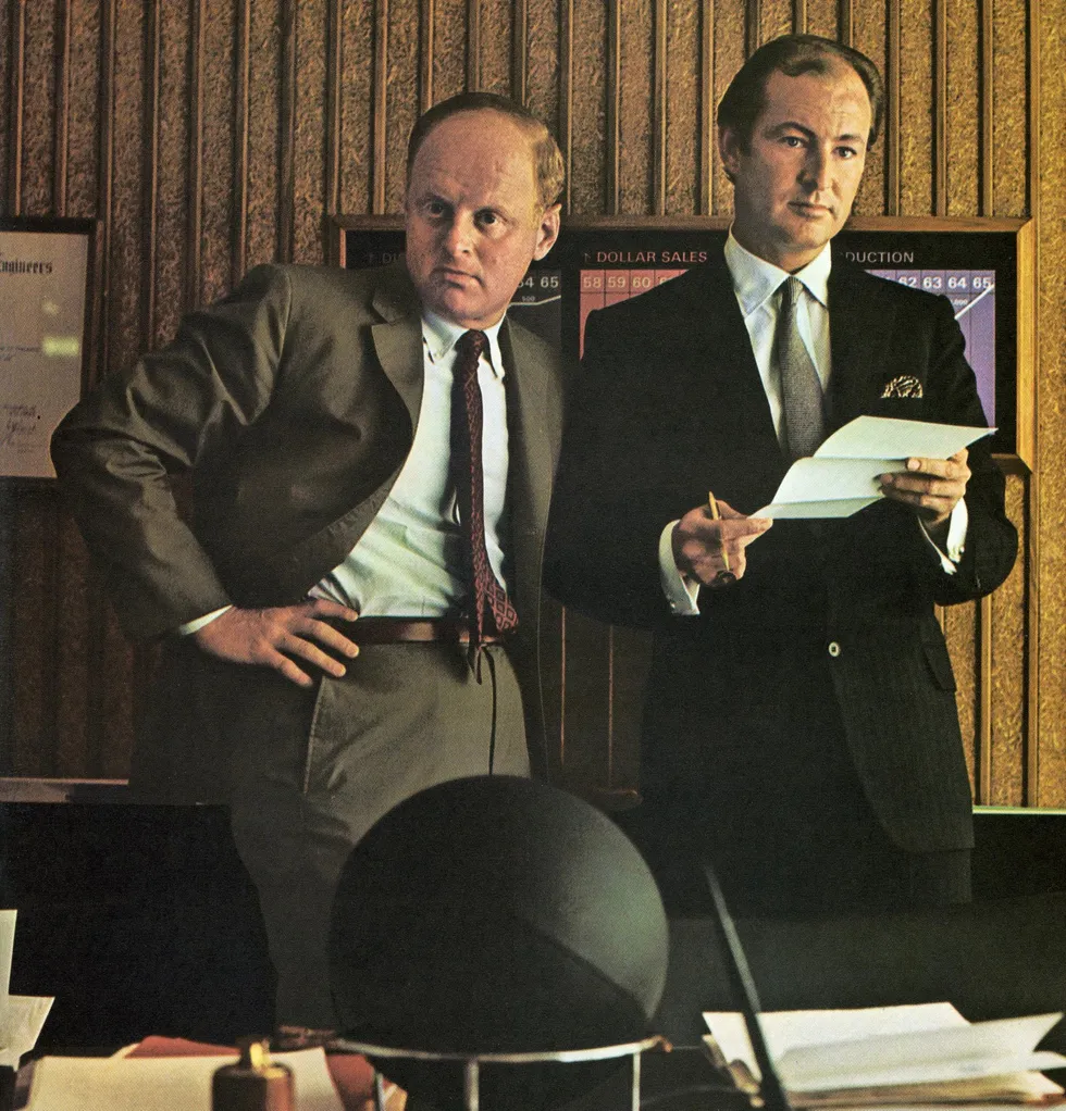 Color photo of two men in office attire. 