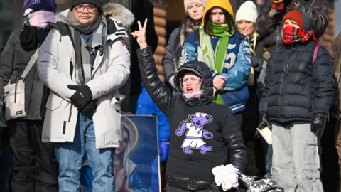 EPA A woman wearing a black puffer jacket with a purple bear motif puts her right arm in the air in a peace sign, at a makeshift memorial for Alex Pretti in south Minneapolis.